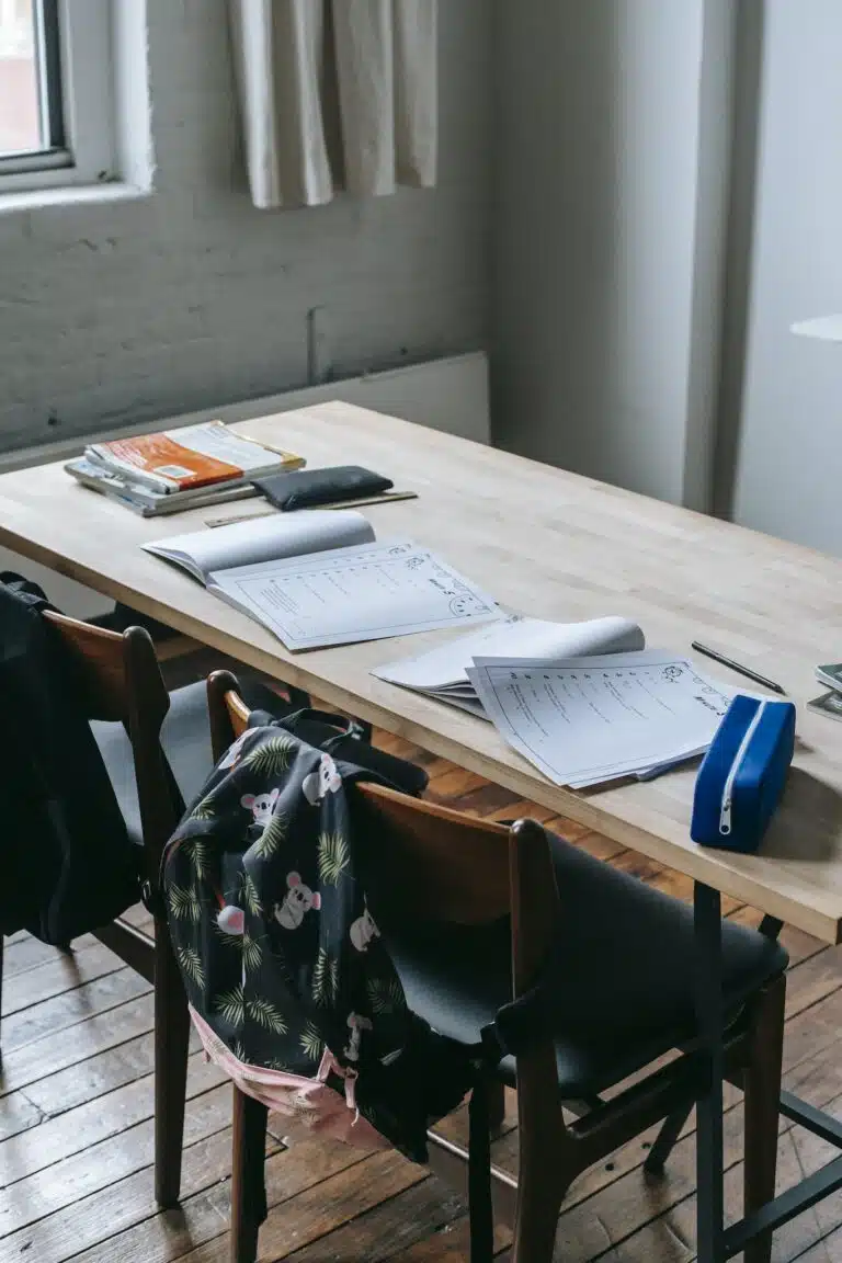 Wooden desk with opened copybooks and workbooks placed near chairs with backpacks in light classroom